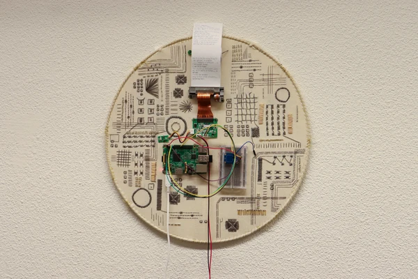 Image of Sadie Girigorie's artwork called a touching interface. Its placed on a wall. It has a round shape with different embroidery. In the center of it is a printer.