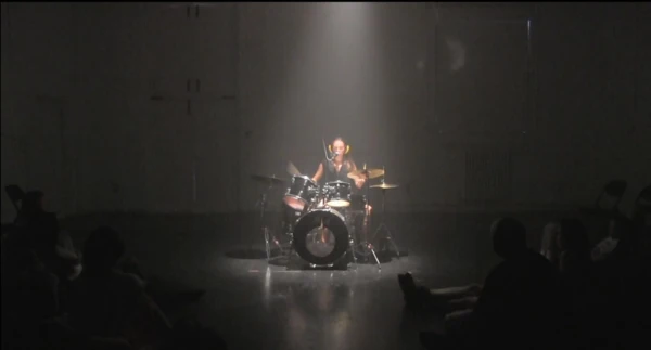 The artist Vida Vojic is setting behind a drum and performing the performance "TIMING IS DIVINE". The photo shows here and the drum in the center with some light focused on her and her setup.