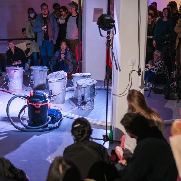 The image contains different metal trash cans and vacuum cleaners. The are centered in the image and highlighted with different lights. Around them you is the audience observing them and listening to them.